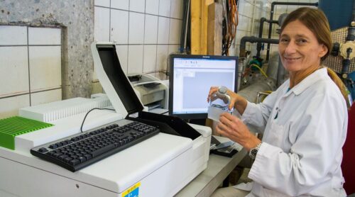 Forscherin im Labor sitzt bei Messgerät und gießt Flüssigkeit aus Flasche in Behälter