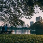 Zwei Menschen sitzen auf Parkbank vor einem See, im Hintergrund die Skyline einer Stadt