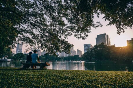 Zwei Menschen sitzen auf Parkbank vor einem See, im Hintergrund die Skyline einer Stadt