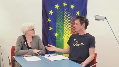 Zwei Menschen diskutieren, im Hintergrund die EU-Flagge, jedoch mit Übergang in bunte Farben.