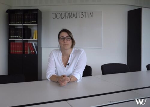 Eine Frau am Konferenztisch blickt in die Kamera. Dahinter ein Whiteboard mit dem Wort "Journalistin".