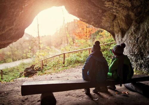 zwei Kinder sitzen unter einem Felsvorsprung auf einer Bank und blicken auf die Landschaft