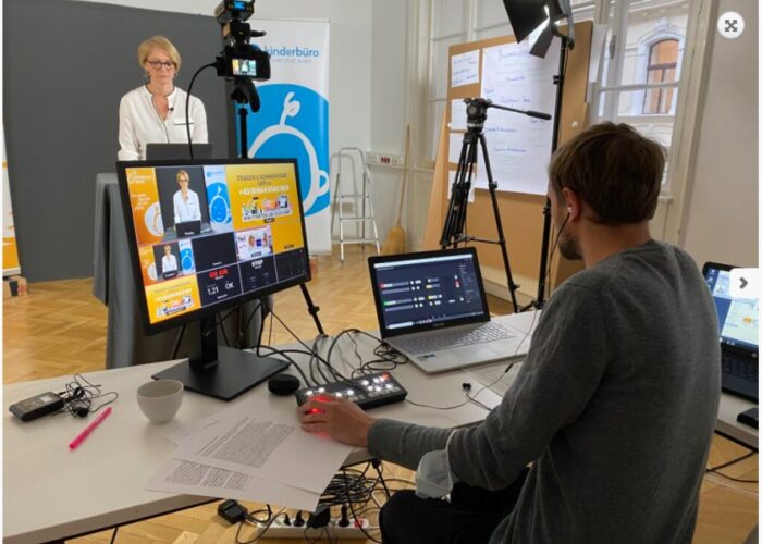 Ein Livestream-Studio mit Kameras, Beleuchtung und zwei Computern. Eine Frau steht vor der Kamera, ein Mann sitzt hinter der Kamera an den Computern.