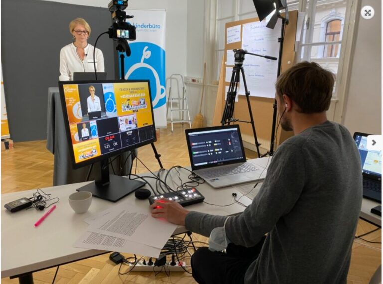 Ein Livestream-Studio mit Kameras, Beleuchtung und zwei Computern. Eine Frau steht vor der Kamera, ein Mann sitzt hinter der Kamera an den Computern.