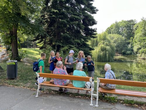 Mehrere Kinder mit Klemmbrettern befragen mehrere ältere Menschen, die auf Parkbänken sitzen.