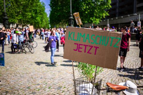 Foto von einer Klimademonstration mit einem Pappschild im Vordergrund, auf dem "Klimaschutz jetzt" steht.