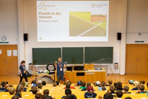 Eine Vorlesung zum Thema "Plastik" in einem großen Uni-Hörsaal. Der Vortragende zeigt den Kindern zwei Plastikteile.