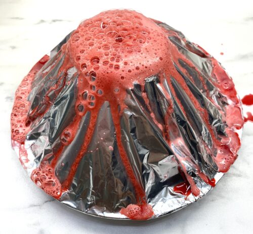 ein vulkanförmiger Kegel aus Alufolie; aus der Öffnung an der Oberseite quillt eine rote Flüssigkeit hervor und rinnt an den Seiten hinunter