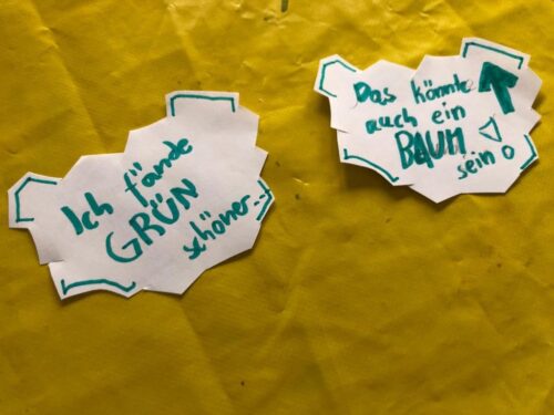 Zwei Zettel liegen auf einem gelben Tischtuch. Auf einem Zettel steht in grünem Filzstift "Ich fände GRÜN schöner", auf dem anderen Zettel steht "Das könnte auch ein Baum sein".