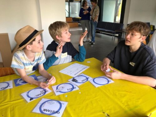 Drei Kinder sitzen am Tisch mit gelbem Tischtuch und diskutieren.