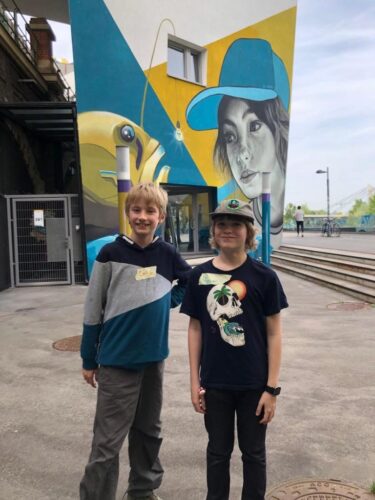 Zwei Kinder stehen vor dem DOCK und schauen in die Kamera. Ein Bub trägt ein schwarzes T-Shirt und eine Kappe, der andere einen blau-grauen Pullover