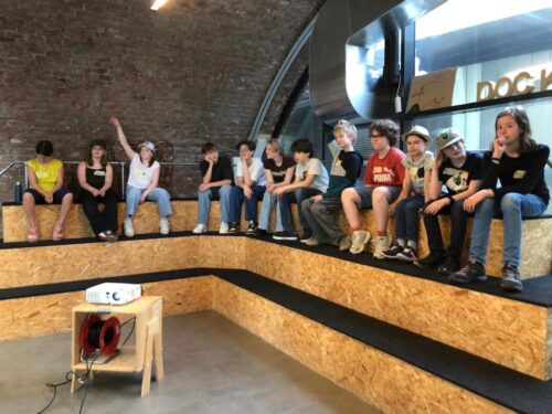 Kinder sitzen auf Tribüne der DOCK-Räumlichkeiten und ein Kind zeigt auf