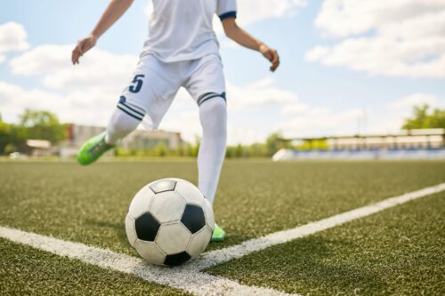 Fußballer mit weißem Dress und grünen Schuhen mit der Nummer 5 ist kurz davor, einen Ball auf einem Rasen zu kicken