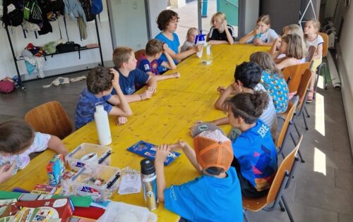 Mehrere Kinder sitzen an einem langen gelben Tisch um eine Vermittlerin herum, die einen Wasserkrug und eine Sprühflasche zeigt