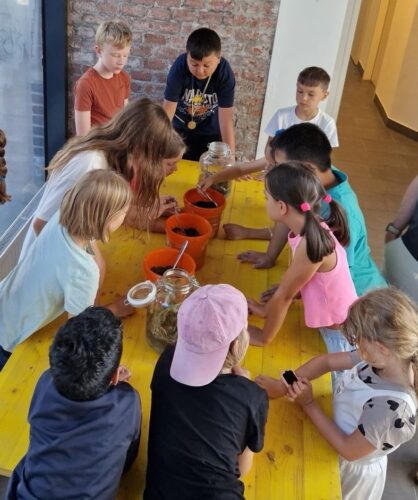 Kinder schauen sich an einem Tisch drei orange Kübel genauer an, die mit verschiedenen Bodenmaterialien befüllt sind