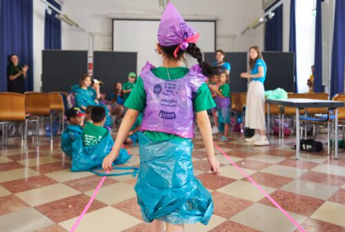 Kind von hinten, das sich mit Plastikteilen gekleidet hat. Andere Kinder sind im Hintergrund zu sehen und applaudieren.