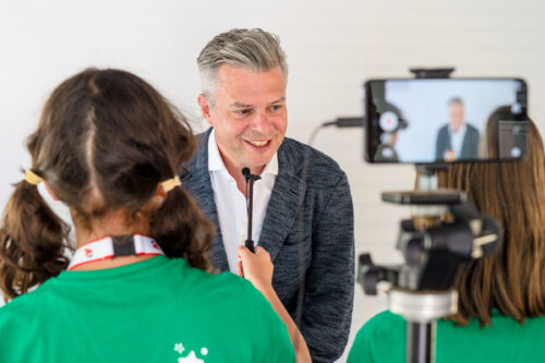 Thomas Arnoldner wird von zwei Mädchen mit einem Mikrofon interviewt. Im Vordergrund sieht man ein Handy auf einem Stativ, das mitfilmt.