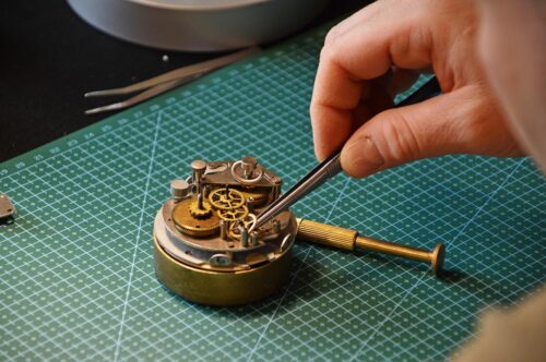 Hand, die mit einer Pinzette ein mechanisches Uhrwerk berührt