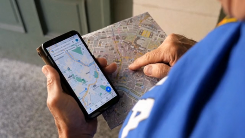 Finger einer Person, die gerade auf einen Stadtplan zeigt und ein Smartphone mit einem Routenplaner in Händen hält