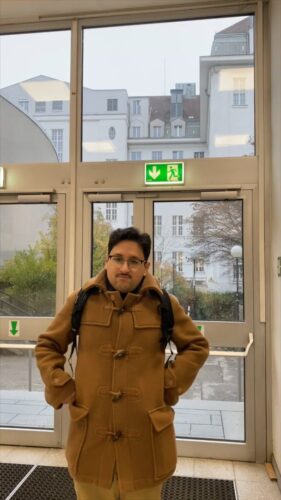 junger Mann in braunem Mantel, der beide Hände an die Hüften hält