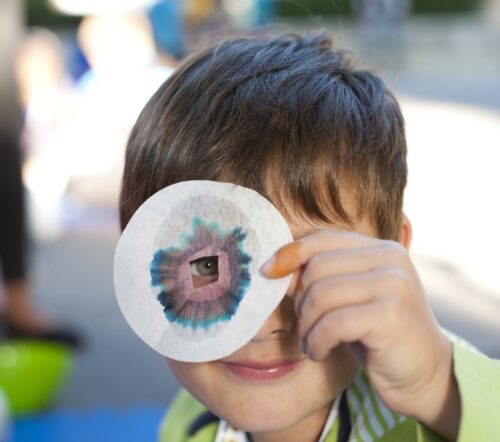 Bub schaut durch ein Filterpapier mit einem Loch in der Mitte, um das herum sich bunte Farbstreifen gebildet haben, weil er das Experiment ausprobiert hat
