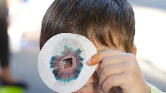 Bub schaut durch ein Filterpapier mit einem Loch in der Mitte, um das herum sich bunte Farbstreifen gebildet haben, weil er das Experiment ausprobiert hat