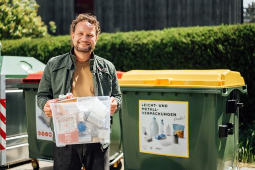 Mann mit einer kleinen Plastikkiste, in der Verpackungen aus Plastik und Metall gesammelt sind. Dahinter steht ein Sammelcontainer mit gelbem Deckel, auf dem "Leicht- und Metallverpackungen" zu lesen ist.