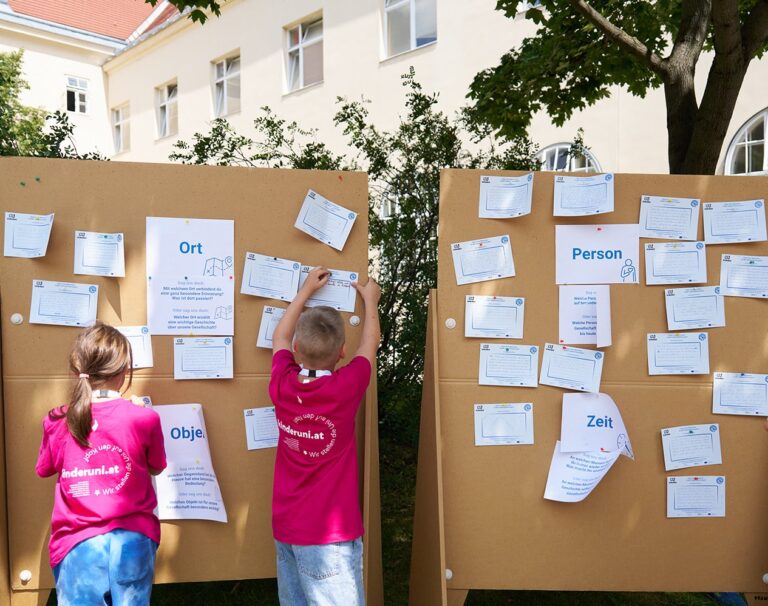 Zwei Kinder pinnen auf dem Universitätscampus einen Zettel an eine Pinnwand, an der bereits mehrere Zettel befestigt sind.